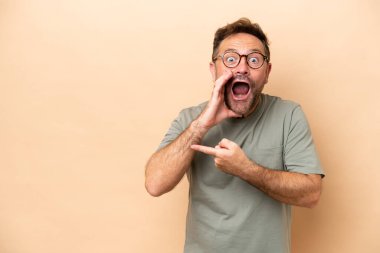 Beyaz tenli, bej arka planda izole edilmiş orta yaşlı bir adam bir ürün sunmak ve bir şeyler fısıldamak için kenarı işaret ediyor.