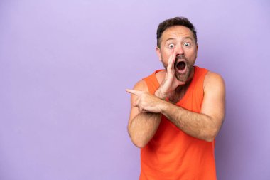 Mor arka planda izole edilmiş orta yaşlı beyaz bir adam bir ürün sunmak ve bir şeyler fısıldamak için kenarı işaret ediyor.