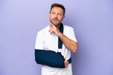 Kırık kollu orta yaşlı beyaz bir adam ve mor arka planda izole edilmiş askı takıyor gülümserken yukarı bakıyor.