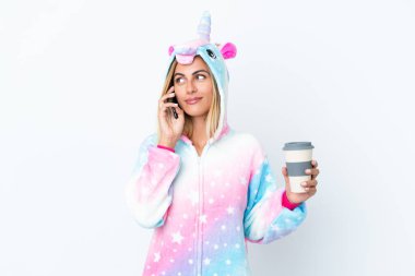 Sarışın Uruguaylı kız tek boynuzlu at pijaması giyiyor. Beyaz arka planda izole edilmiş. Elinde kahve ve cep telefonu var.