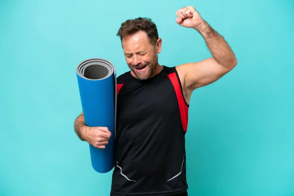 Middle age sport man going to yoga classes while holding a mat isolated on blue background celebrating a victory