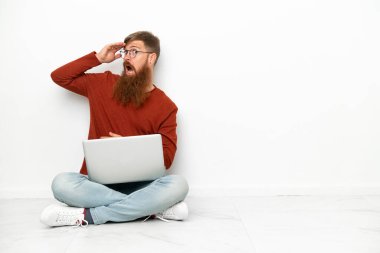 Beyaz arka planda dizüstü bilgisayarı olan genç, beyaz beyaz bir adam yan tarafa bakarken sürpriz jest yapıyor.