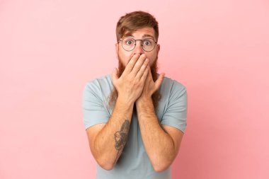 Kırmızımsı beyaz adam pembe arka planda izole edilmiş ağzı elleriyle kapatıyor.