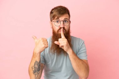Pembe arka planda izole edilmiş genç, kızıl beyaz bir adam. Yan tarafı işaret ediyor ve sessizlik hareketi yapıyor.