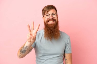 Pembe arka planda izole edilmiş genç beyaz adam mutlu ve parmaklarıyla üçe kadar sayıyor.