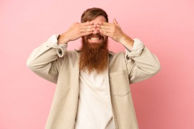 Kırmızımsı beyaz adam pembe arka planda izole edilmiş gözlerini elleriyle kapatmış ve gülümsüyor.