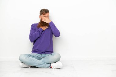 Beyaz arka planda izole bir şekilde yerde oturan Kafkasyalı genç adam öksürük ve kötü hissetmekten muzdarip.