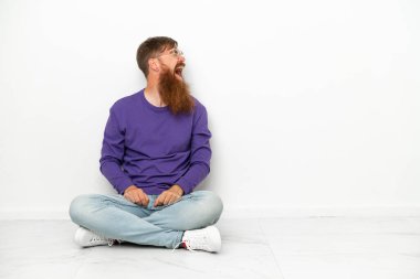 Beyaz arka planda izole bir şekilde yerde oturan Kafkasyalı genç adam yan yana gülüyor.