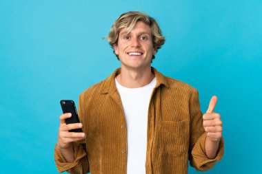 İngiliz adam mavi arkaplan üzerinde kapalı cep telefonu kullanarak başparmağını kaldırıyor