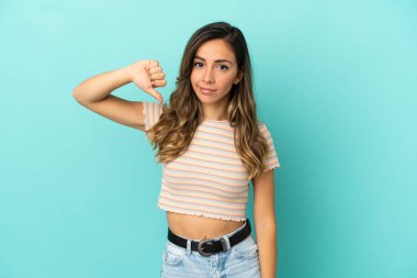 İzole edilmiş mavi arka planda genç bir kadın baş parmağını negatif ifadeyle aşağı gösteriyor.