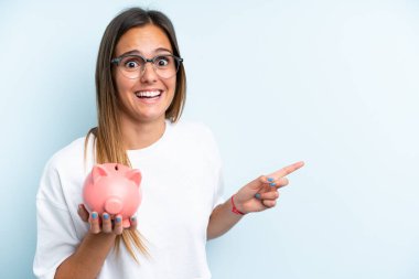 Young caucasian woman holding a piggybank isolated on blue background surprised and pointing finger to the side