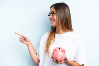 Mavi arka planda izole edilmiş domuz kumbarası tutan beyaz bir kadın bir ürünü sunmak için kenarı işaret ediyor.