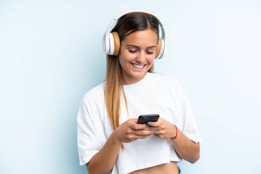 Genç beyaz kadın mavi arka planda izole edilmiş müzik dinliyor ve cep telefonu arıyor.