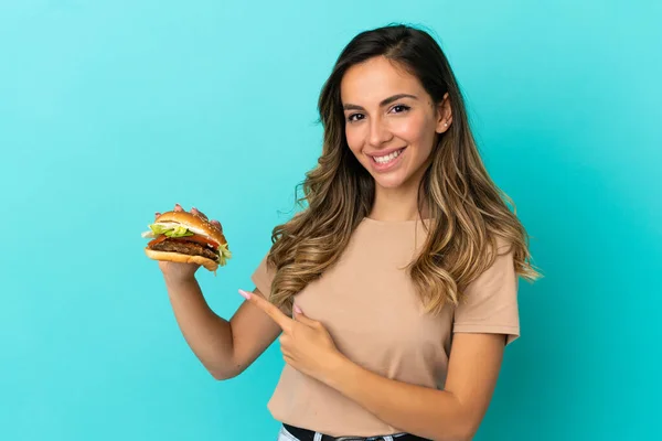 Młoda Kobieta Trzyma Hamburgera Odosobnionym Tle Wskazując Bok Aby Przedstawić — Zdjęcie stockowe