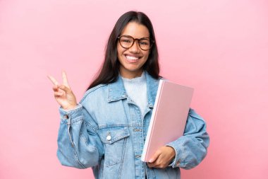 Kolombiyalı genç bir kız pembe arka planda izole edilmiş gülümsüyor ve zafer işareti gösteriyor.