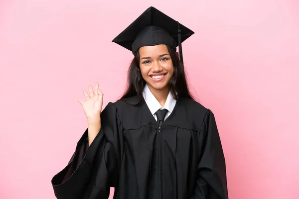 Young university Colombian woman graduate isolated on pink background saluting with hand with happy expression