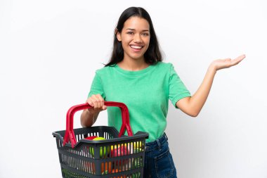 Young Colombian woman holding a shopping basket full of food isolated on white background extending hands to the side for inviting to come