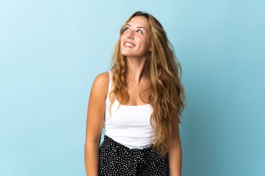 Mavi arka planda izole edilmiş genç sarışın kadın yukarı bakarken bir fikir düşünüyor.