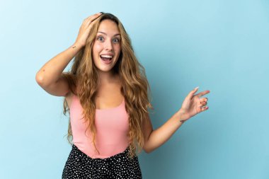 Mavi arka planda izole edilmiş genç sarışın kadın şaşırmış ve işaret ediyor.