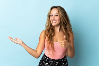 Mavi arka planda izole edilmiş genç sarışın bir kadın. Avucunun içinde hayali bir şey tutuyor. Bir ilan vermek için.