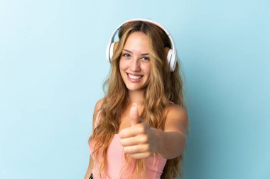 Young blonde woman isolated on blue background listening music and with thumb up