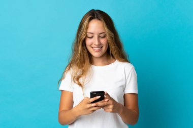 Mavi arka planda izole edilmiş genç sarışın kadın cep telefonuyla bir mesaj gönderiyor.