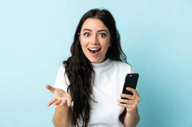Mavi arka planda izole edilmiş cep telefonu kullanan genç bir kadın.