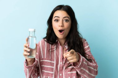 Mavi arka planda su izole edilmiş genç bir kadın şaşırmış ve ön tarafı işaret ediyor.