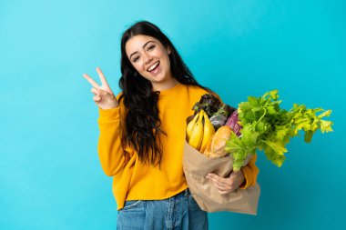 Elinde alışveriş çantası tutan genç bir kadın mavi arka planda izole edilmiş gülümsüyor ve zafer işareti gösteriyor.