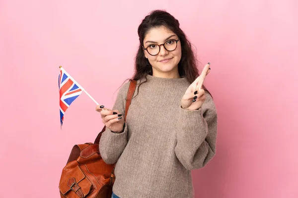 Young Russian woman holding an United Kingdom flag isolated on pink background with fingers crossing and wishing the best