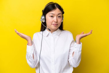 Tele pazarlamacı Çinli kadın sarı arka planda izole edilmiş bir kulaklıkla çalışıyor. El kaldırırken şüpheleri var.