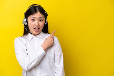 Sarı arka planda izole edilmiş kulaklıkla çalışan pazarlamacı Çinli kadın şaşırmış ve işaret ediyor.
