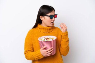 Young Chinese woman isolated on white background with 3d glasses and holding a big bucket of popcorns while looking side
