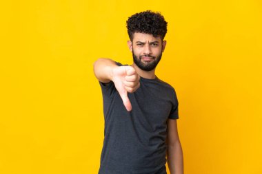Sarı arka planda izole edilmiş genç Faslı adam olumsuz ifadeyle baş parmağını aşağı gösteriyor.