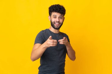 Sarı arka planda izole edilmiş genç Faslı adam şaşırmış ve ön tarafı işaret ediyor.