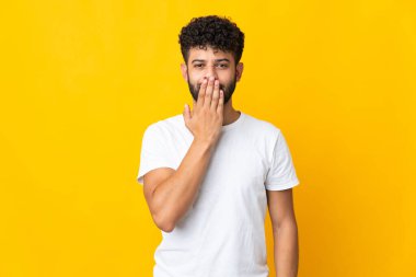 Faslı genç bir adam sarı arka planda izole edilmiş. Ağzı eliyle kapatıyor.