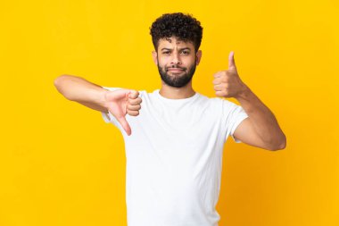 Genç Faslı adam sarı arka planda izole edilmiş, iyiye kötüye işaret yapıyor. Evet ya da hayır arasında kararsız