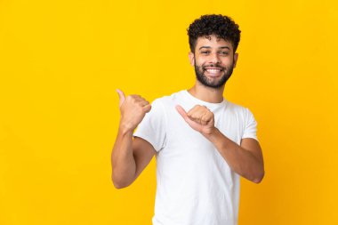 Sarı arka planda izole edilmiş genç Faslı bir adam bir ürün sunmak için kenarı işaret ediyor.