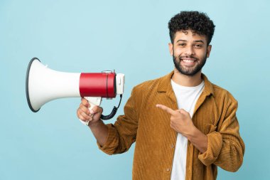 Young Moroccan man isolated on blue background holding a megaphone and pointing side