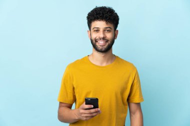 Genç Faslı adam mavi arka planda cep telefonu kullanarak izole edildi