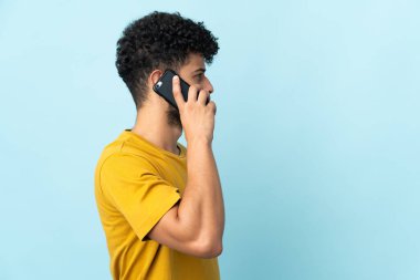 Faslı genç bir adam mavi arka planda izole edilmiş ve cep telefonuyla sohbet ediyor.