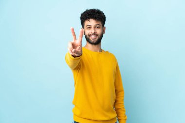 Mavi arka planda izole edilmiş genç Faslı adam gülümsüyor ve zafer işareti gösteriyor.