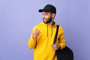 Mor arka planda izole edilmiş spor çantalı genç Faslı bir adam bir ürünü sunmak için kenarı işaret ediyor.