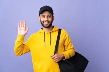 Spor çantalı genç Faslı adam mor arka planda izole edilmiş mutlu bir ifadeyle selam veriyor.