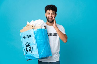 Genç Faslı bir adam, izole edilmiş bir arkaplanı şaşırmış ve şok olmuş bir yüz ifadesiyle geri dönüştürmek için elinde kağıt dolu bir geri dönüşüm çantası tutuyor.