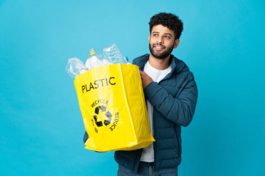 Bir çanta dolusu plastik şişeyi elinde tutan genç Faslı adam izole edilmiş arka planda geri dönüşüm yapmak için yukarı bakıp gülümsüyor.