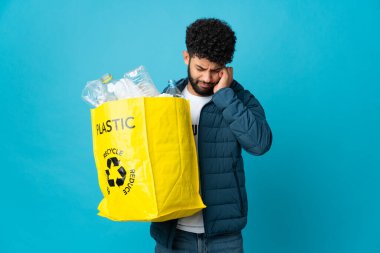 Genç Faslı bir adam, izole edilmiş arka plan üzerinde geri dönüşüm yapmak için elinde plastik şişelerle dolu bir çanta tutarken hayal kırıklığına uğramış ve kulaklarını kapatmış.