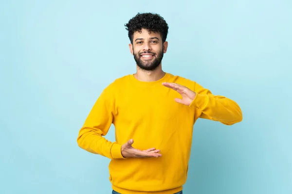 Young Moroccan man isolated on blue background holding copyspace imaginary on the palm to insert an ad