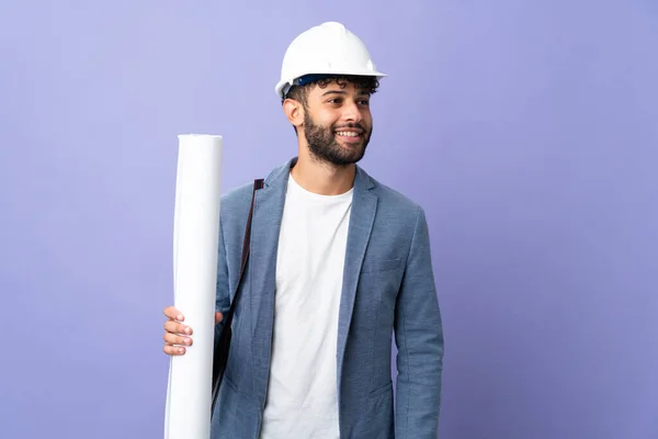 Young architect Moroccan man with helmet and holding blueprints over isolated background posing with arms at hip and smiling