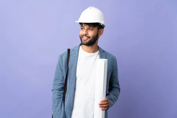 Young architect Moroccan man with helmet and holding blueprints over isolated background looking to the side and smiling